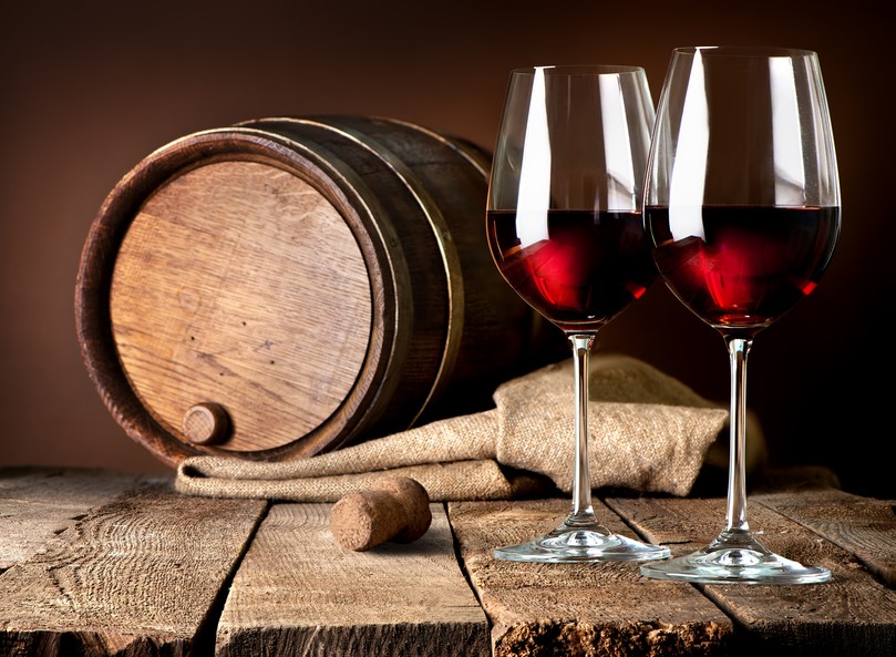 two glasses of red wine next to a wine barrel and a cork